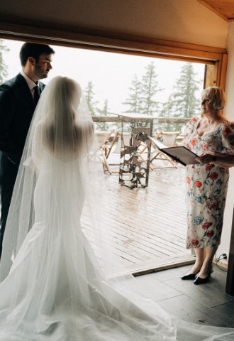 Wedding couple standing together in a mountain cabin overlooking the forest in Switzerland. Officiant is speaking to them. At the side table there is a candle ready for a candle lighting ritual.