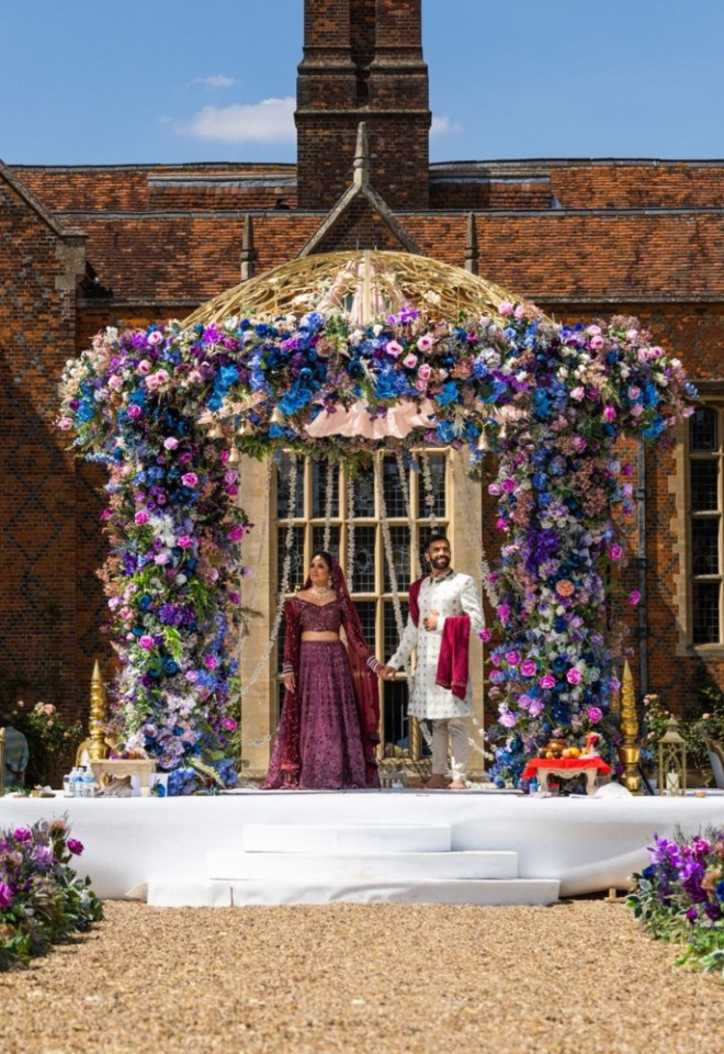 Bride and groom standing under a gold dome mandap covered in vibrant purple and blue flowers on the lawn at North Mymms Park, set against the historic red-brick mansion house