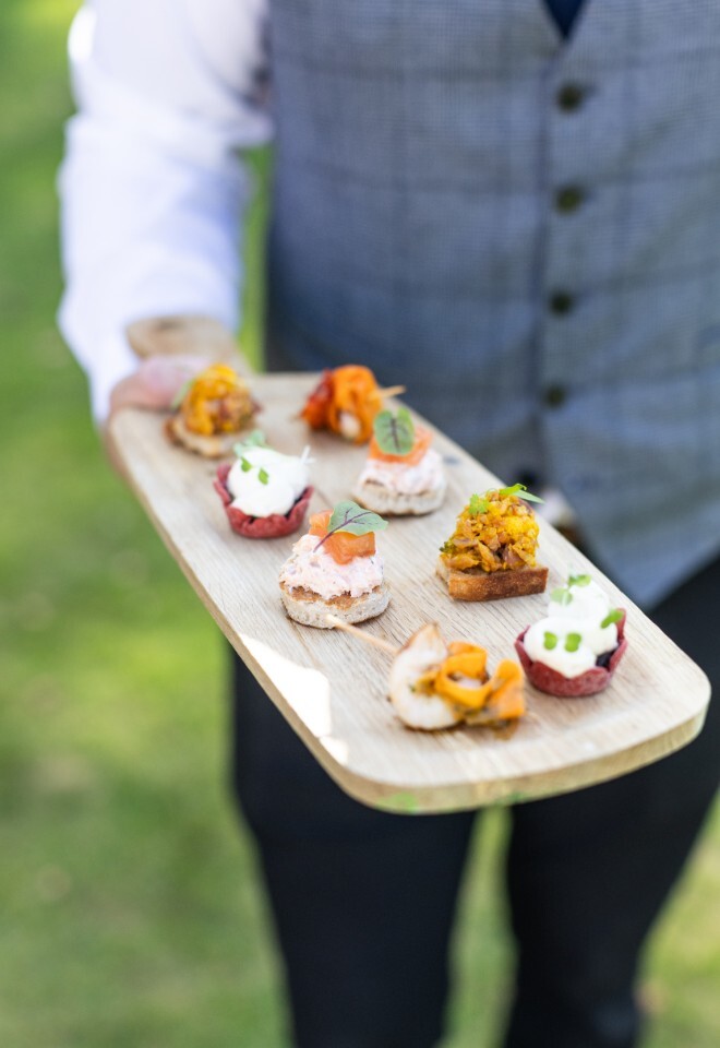 A waiter in a grey waistcoat holds a wooden board of colourful canapés, beautifully arranged and garnished, served outdoors at Washingborough Hall Hotel.