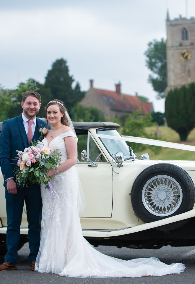 Happy bride and groom in front of their vintage wedding car with church in the far distance