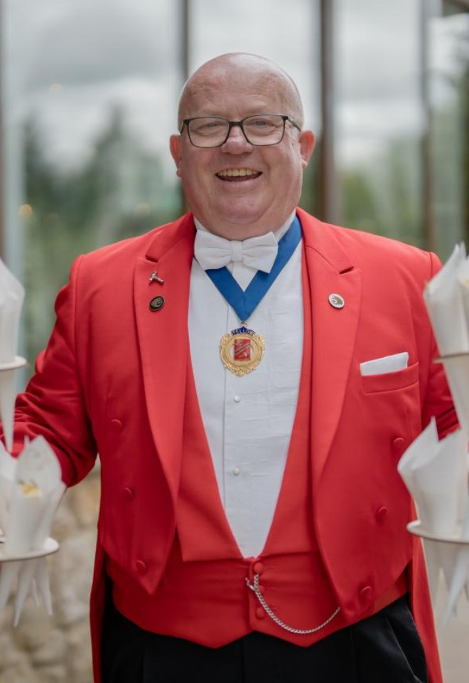 Surrey wedding toastmaster and celebrant Toastmaster John smiling in his traditional red tailcoat while preparing confetti cones for a wedding celebration.