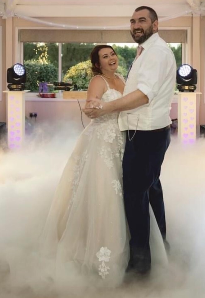 A smiling bride and groom holding hands during their first dance on a floor covered in thick, low-lying fog, flanked by indoor sparkler fountains and DJ lighting equipment with a "Lux Vita Photography