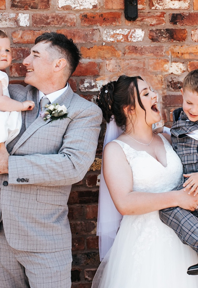 Bride and groom sharing a playful family moment with two young children against the rustic brickwork at The Cock O’Barton.