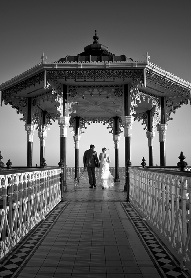 Bride and groom walking away from camera with dog on lead on small pier by the Grand Hotel in Brighton. Black and white wedding photography