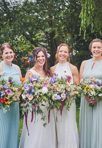 brides in white dresses, bridesmaids in sage green dresses, groomsmen stood posting with summer flower bouquets. Married at Higher Holcombe outdoor wedding venue in Devon. 