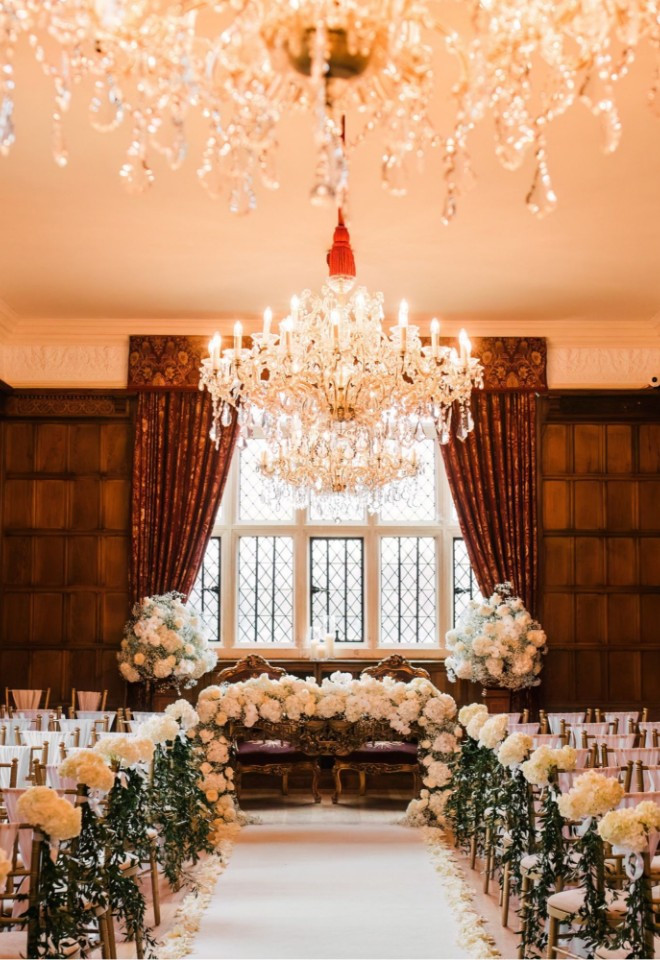 Ceremony setup in the Jacobean Dining Room at North Mymms Park, with a white carpet aisle, floral decor, gold chairs, and crystal chandeliers.