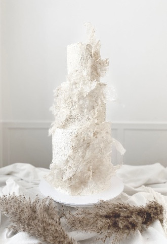 White three tier abstract art cake decorated with rice paper textured buttercream on a white cake stand with pampas branches in the forefront and a neutral toned background 