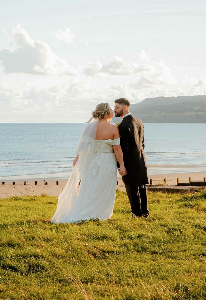 Bride and groom holding hands on a grassy hill overlooking the sea
