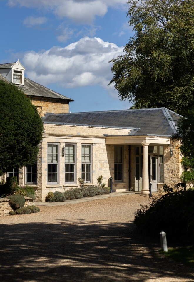 The Orangery at Washingborough Hall Hotel, a light-stone extension with tall windows, columns, and a gravel drive framed by trees under a bright blue sky.