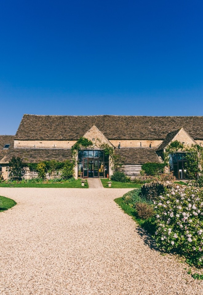 Exterior of Great Tythe Barn with a long Cotswold stone façade, central glass entrance, gravel driveway, and landscaped gardens under a clear blue sky