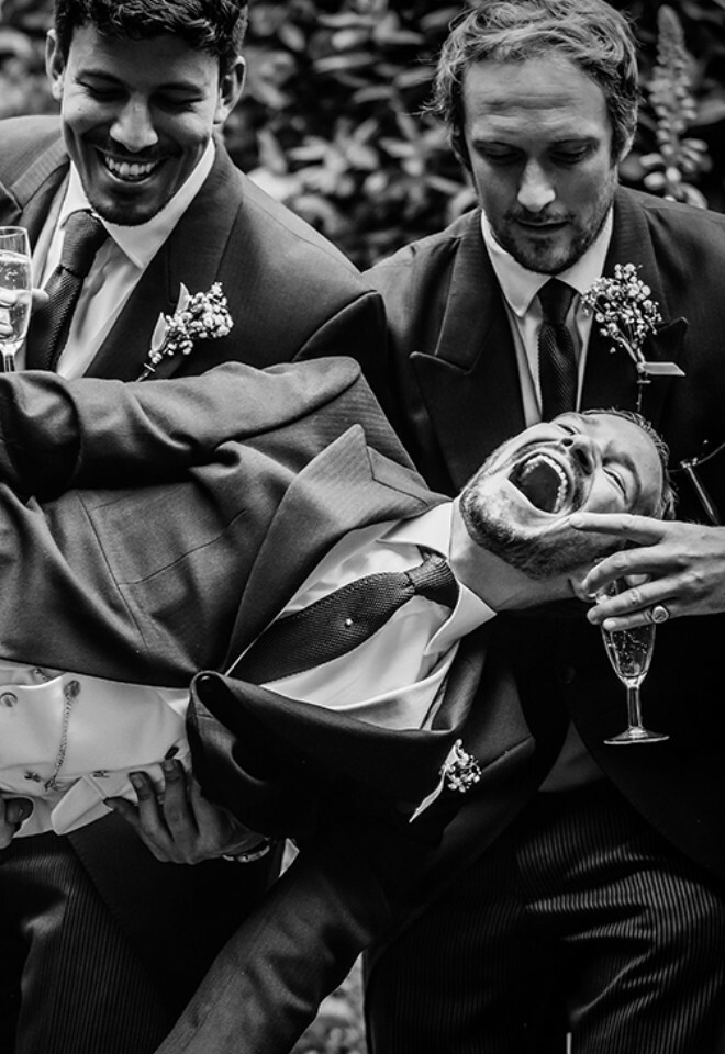 Groom laughing as his groomsmen lift him in the air during a fun wedding moment – black and white candid wedding photography.