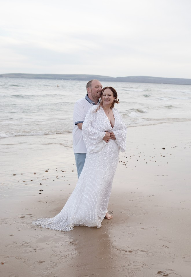 The bride and groom embrace in a bear hug on the sand with the waves crashing behind them, both are smiling and watching their children play just out of shot. 