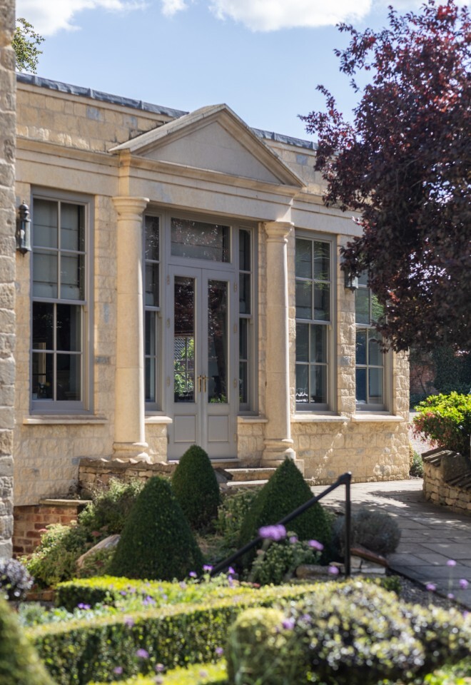 Elegant stone building at Washingborough Hall Hotel with tall windows, columns, and manicured gardens in the foreground, framed by trees under a bright sky.