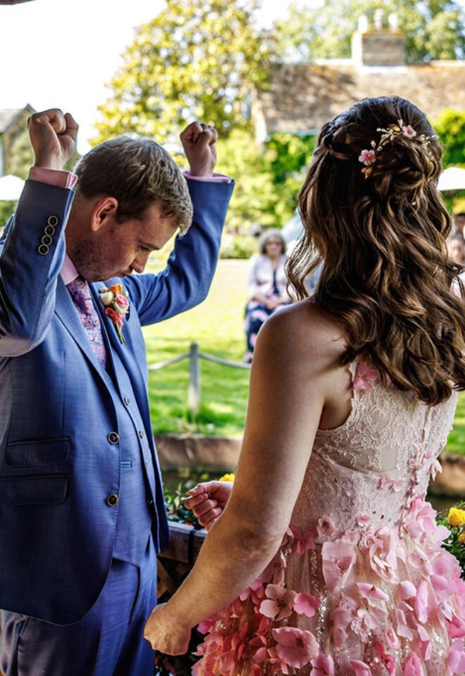 A groom celebrates as hes standing at his wedding ceremony with a bride wearing ornate pink dress