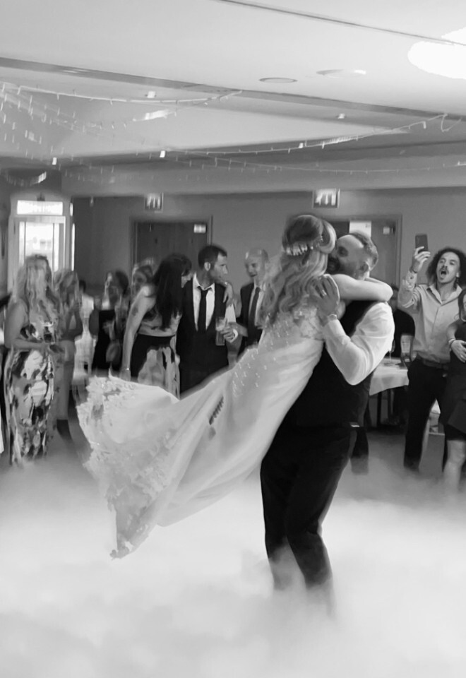 A black and white photo of a groom lifting and spinning his bride on a fog-covered dance floor during their first dance, surrounded by cheering wedding guests, photographers, and videographers.