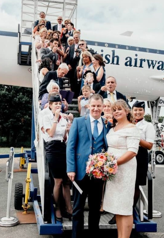 An aviation-themed wedding group photo featuring a bride and groom posing at the bottom of the boarding stairs of the historic Concorde airplane at Brooklands Museum, with wedding guests.