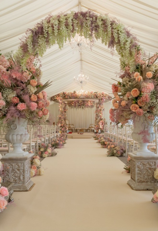 Mandap set up for a Hindu wedding ceremony inside the Pavilion at North Mymms Park, featuring a pastel pink floral arch entrance, stone Ganesha statues, a white carpeted aisle, and a draped mandap at 