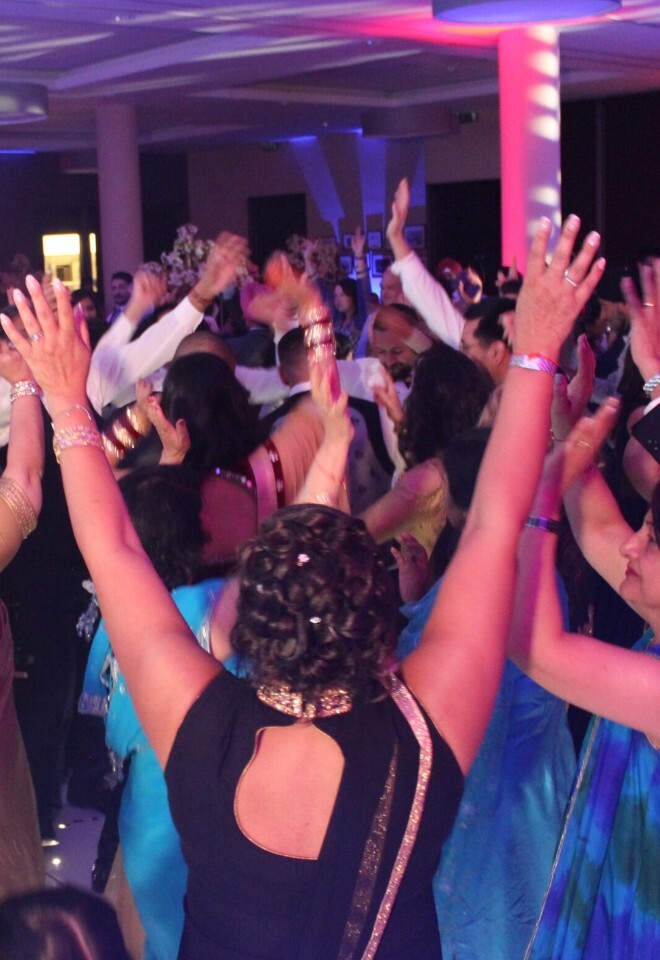 A large crowd of wedding guests, many in traditional South Asian attire, dancing enthusiastically with their hands raised at a lively reception party under vibrant event lighting.