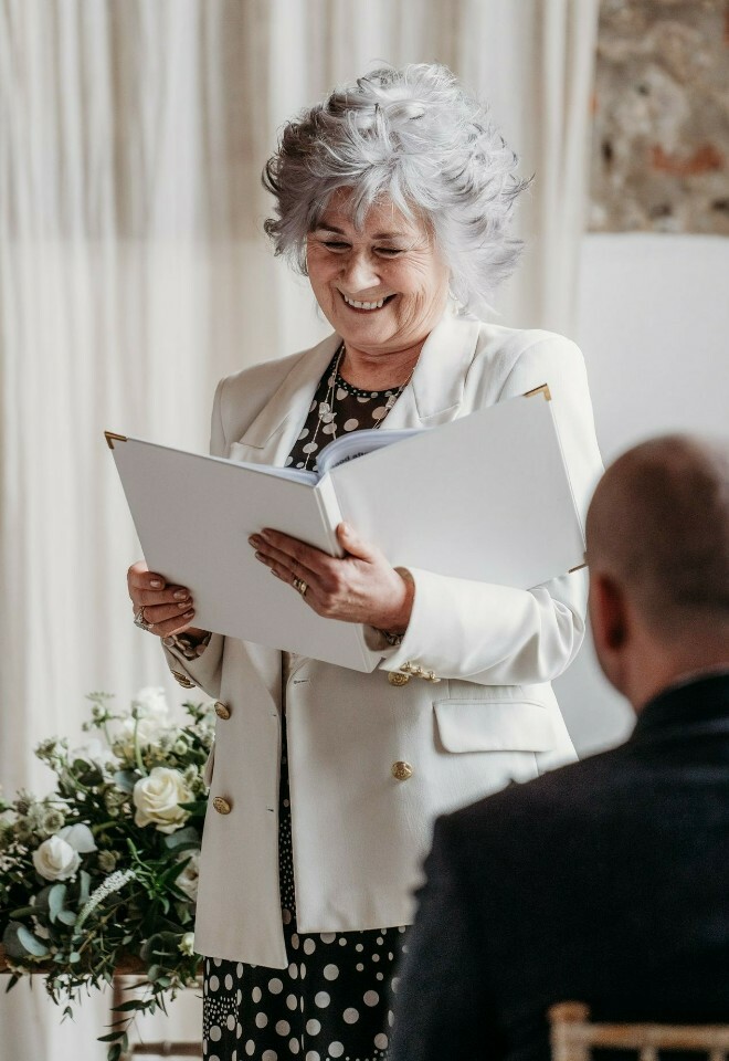 Norfolk celebrant smiling dressed in a white jacket and black and white spotted dress holding a white ceremony folder during the ceremony