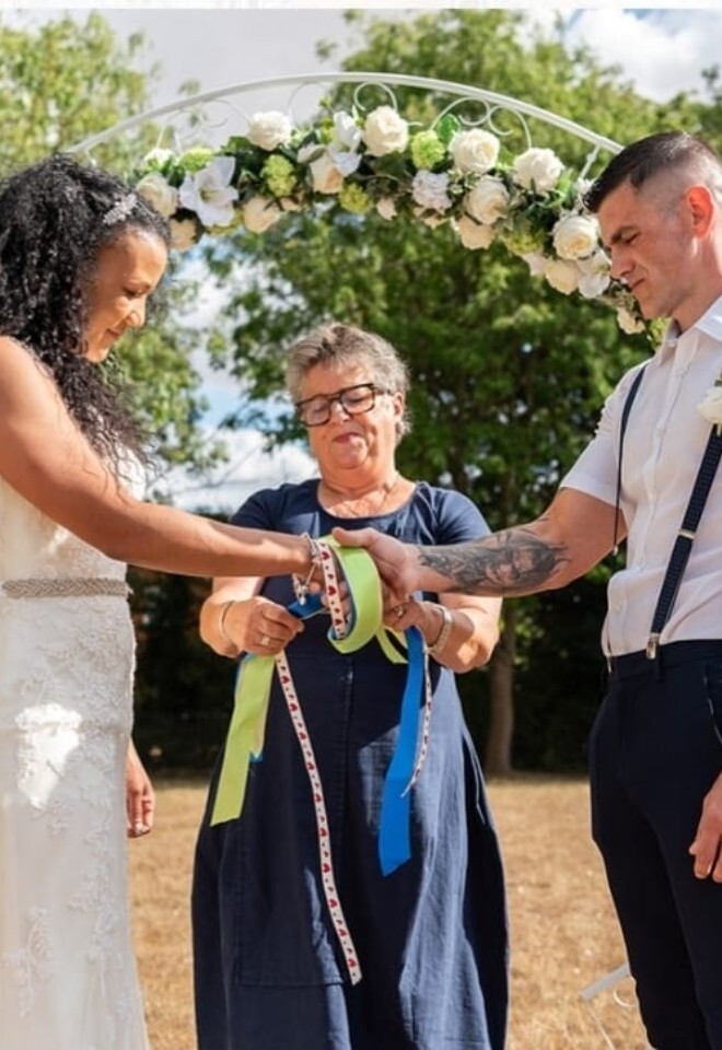 Outdoor wedding ceremony beneath a floral arch with celebrant Dawn Rees guiding a handfasting ritual for the couple