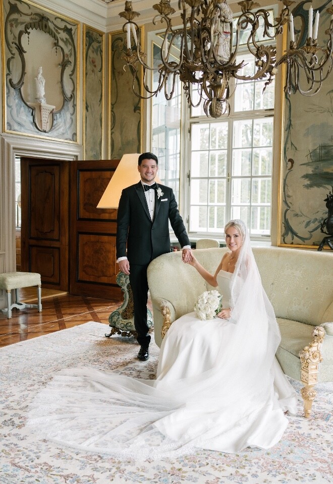 Schloss Leopoldskron Salzburg is where the bride and groom chose to marry. They are in the salon of the palace. The bide in white is seated while the groom in a black tuxedo is standing holding hands.