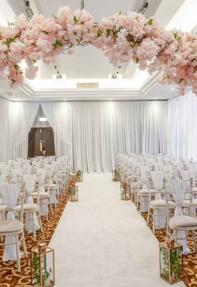 Indoor wedding ceremony set-up at Grand Hotel Gosforth Park with blossom arch, aisle lanterns and white chairs.