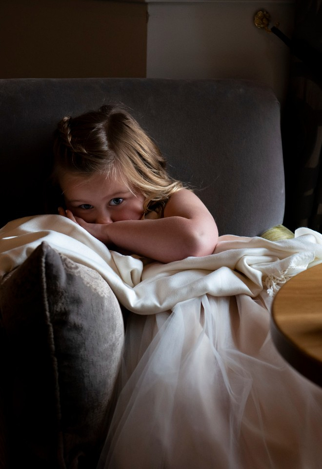 a young flower girl curls up shyly in a chair at a family wedding, lit by a small amount of sunlight streaming in at the nearby window