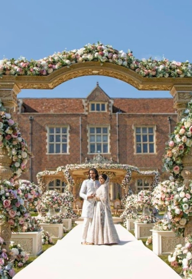 Bride and groom standing on a white aisle framed by an ornate floral arch, with an outdoor mandap setup and the red-brick mansion at North Mymms Park in the background under a blue sky.