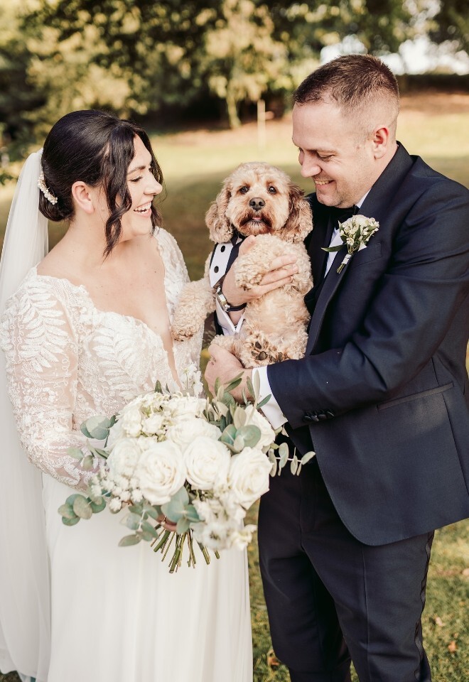 Bride and groom holding their dog dressed in a tuxedo, surrounded by greenery. Image by Sam Rundle Photography.