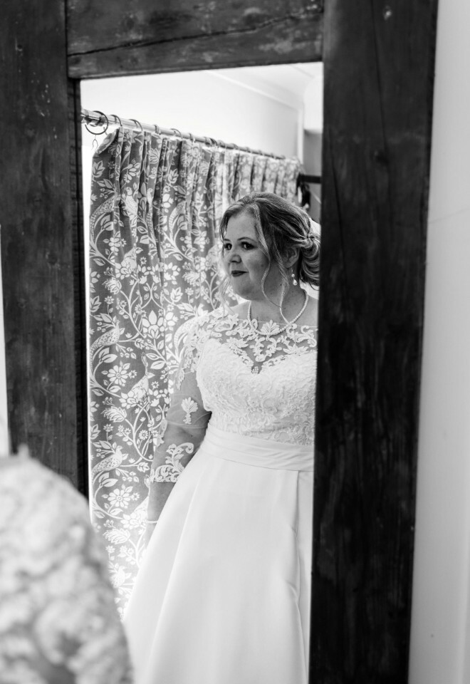 Bride in lace dress looking at her reflection in a rustic mirror, black and white photo.