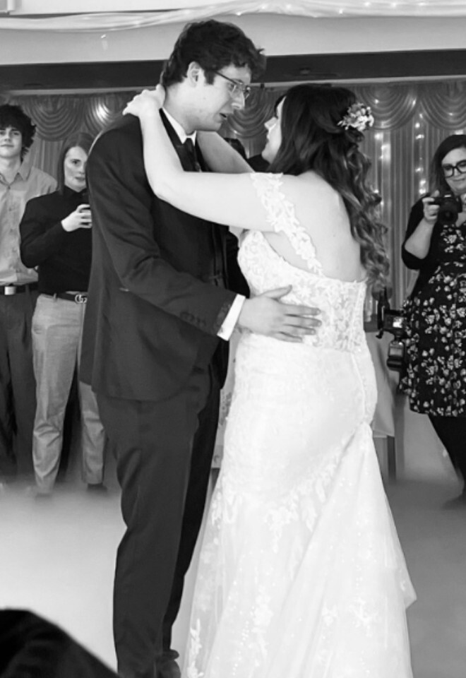 A black and white photo of a bride and groom embracing during their first dance on a fog-covered floor, flanked by indoor sparkler fountains with wedding guests watching under a vaulted ceiling draped