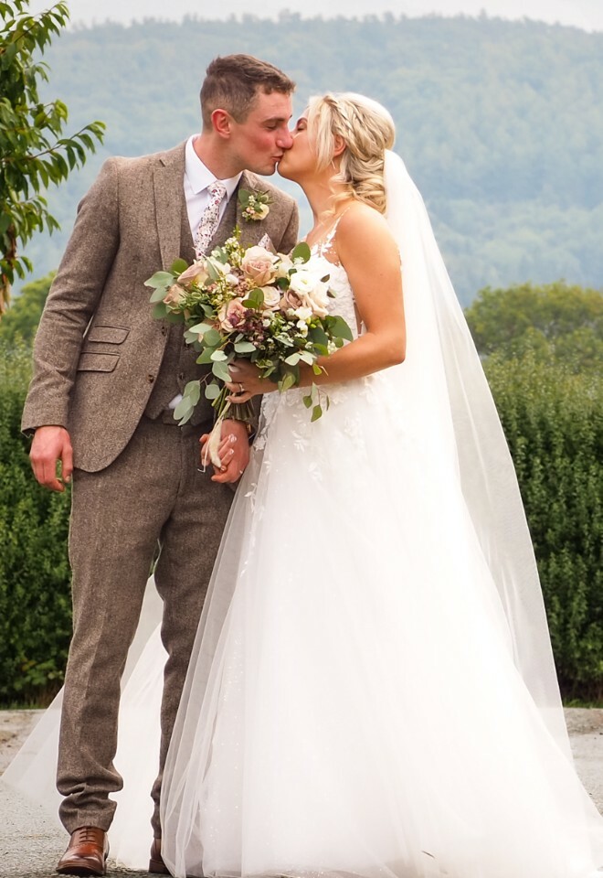 A couple shares a romantic moment on a country road with rolling hills and trees in the background on their wedding day.
