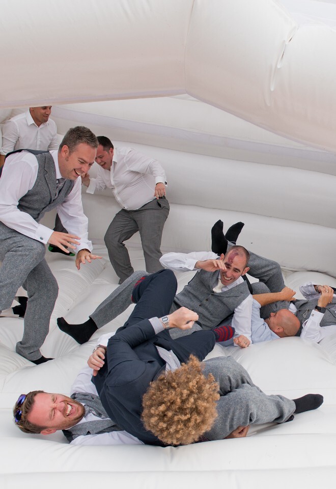 Groom and groomsmen fall over laughing on a white bouncy castle in the gardens of Trunkwell House