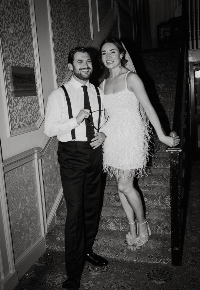 Black-and-white photo of a smiling couple on a staircase at Washingborough Hall Hotel, with the bride in a feathered dress and the groom in suspenders holding sunglasses.