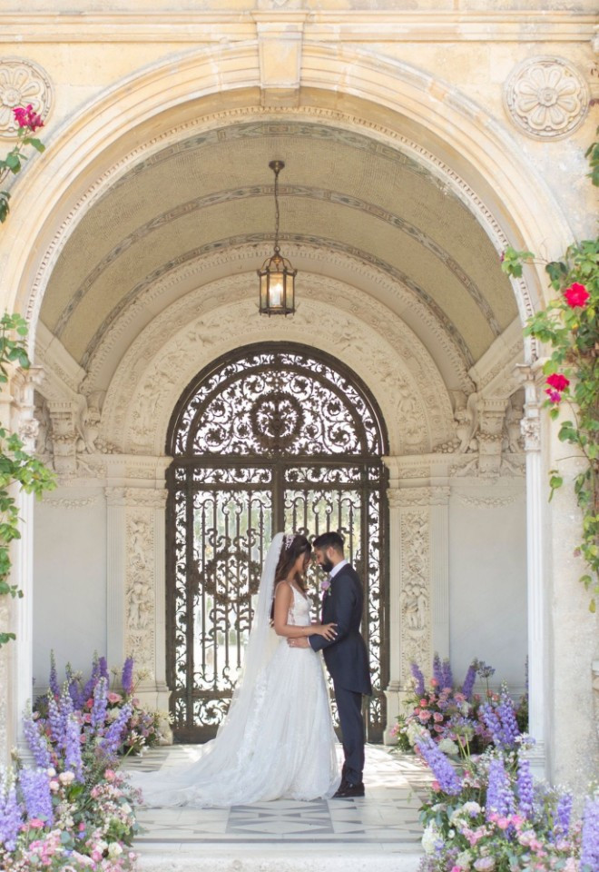 Bride and groom standing together inside the Loggia at North Mymms Park during a civil ceremony, set against stone arches, intricate wrought-iron gates, and lush floral displays.