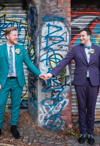 Two grooms hold hands and smile at one another in front of graffiti covered garages