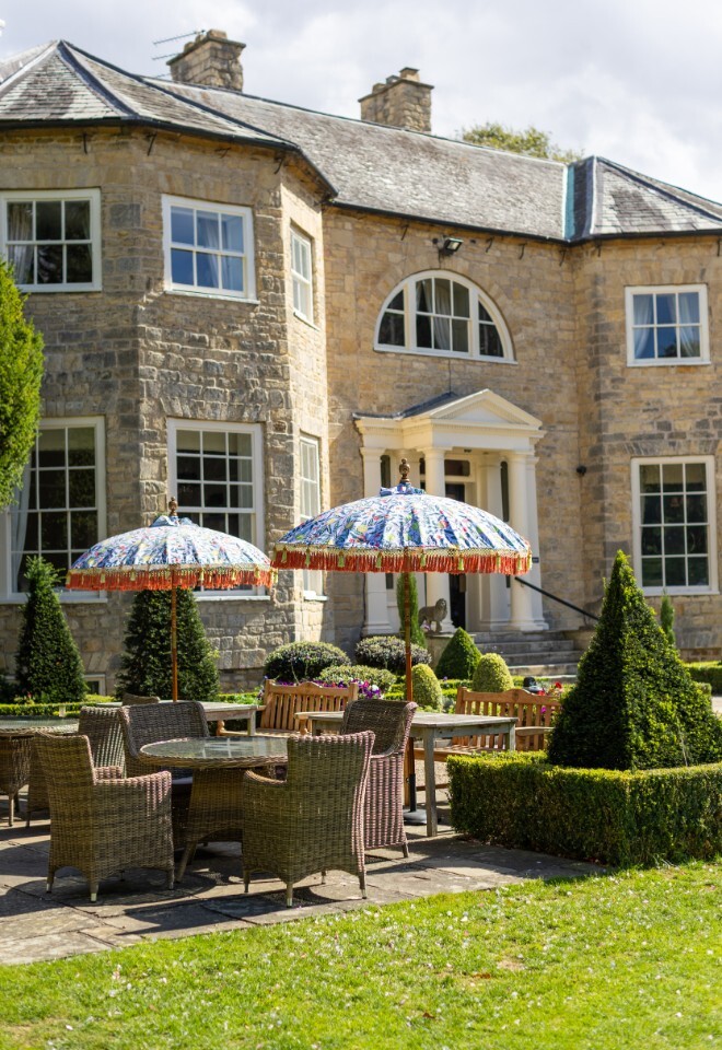 Outdoor seating at Washingborough Hall Hotel, featuring wicker chairs, colourful parasols, and manicured gardens against the elegant Georgian stone façade.