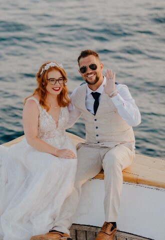 bride and groom smile and wave to the camera as they sit at the end on a yacht at their wedding reception in cyprus