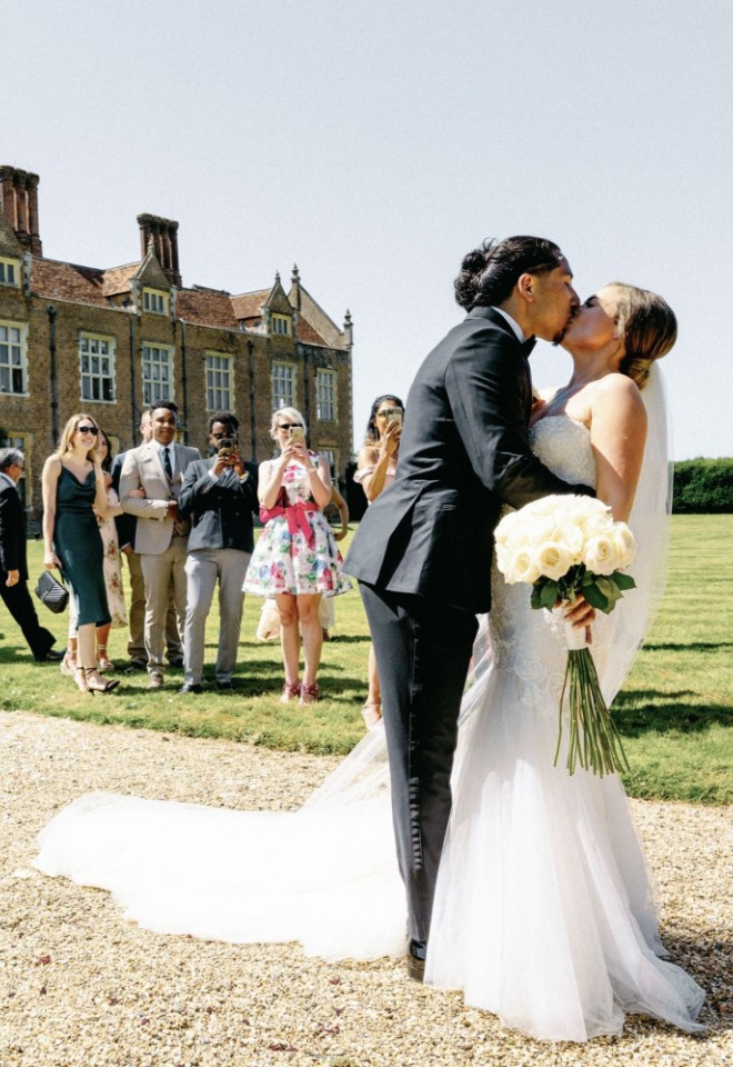 Newlywed couple kissing on the lawn at North Mymms Park, with wedding guests gathered behind them and the historic manor house in the background.