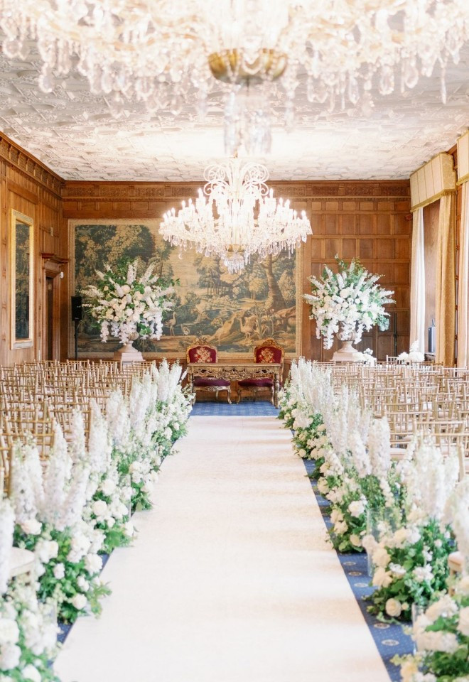 Civil ceremony setup in the Long Gallery at North Mymms Park, featuring a white carpet aisle lined with lush white flowers, gold Chiavari chairs, ornate crystal chandeliers, and wood-paneled walls ado