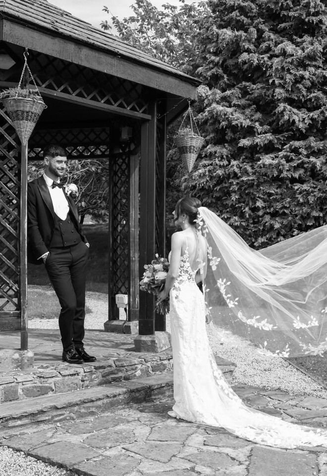 groom stood in pergola looking at the bride whos veil is flying in the wind at Mytton Fold