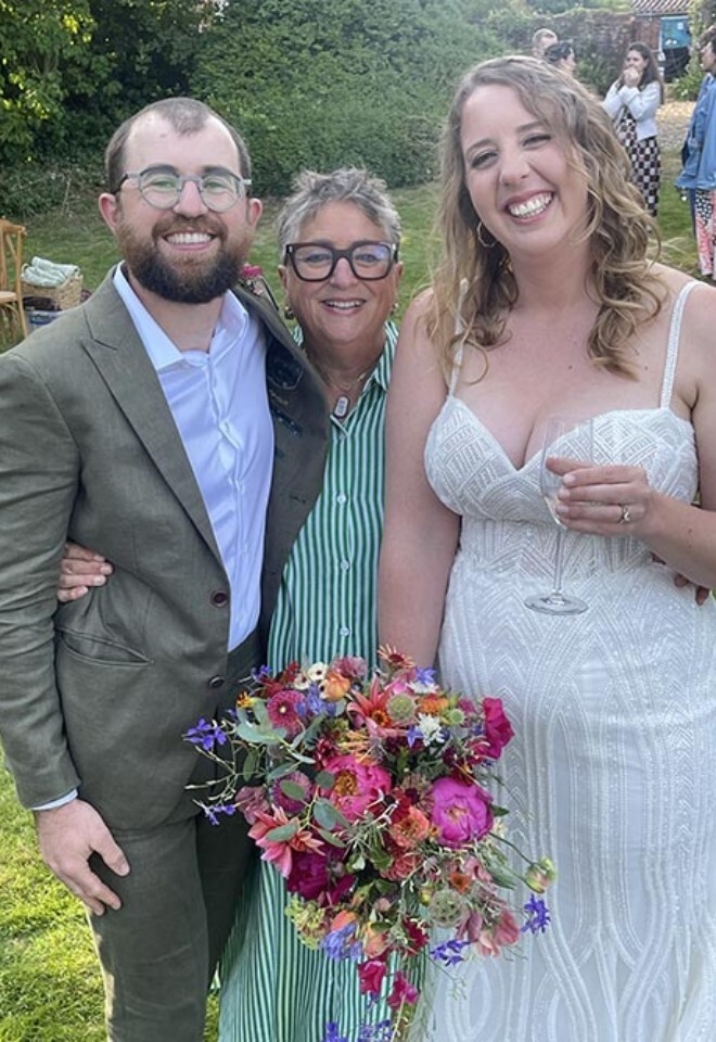 Wedding couple smiling with celebrant Dawn Rees outdoors, holding a colourful bouquet during a garden wedding reception