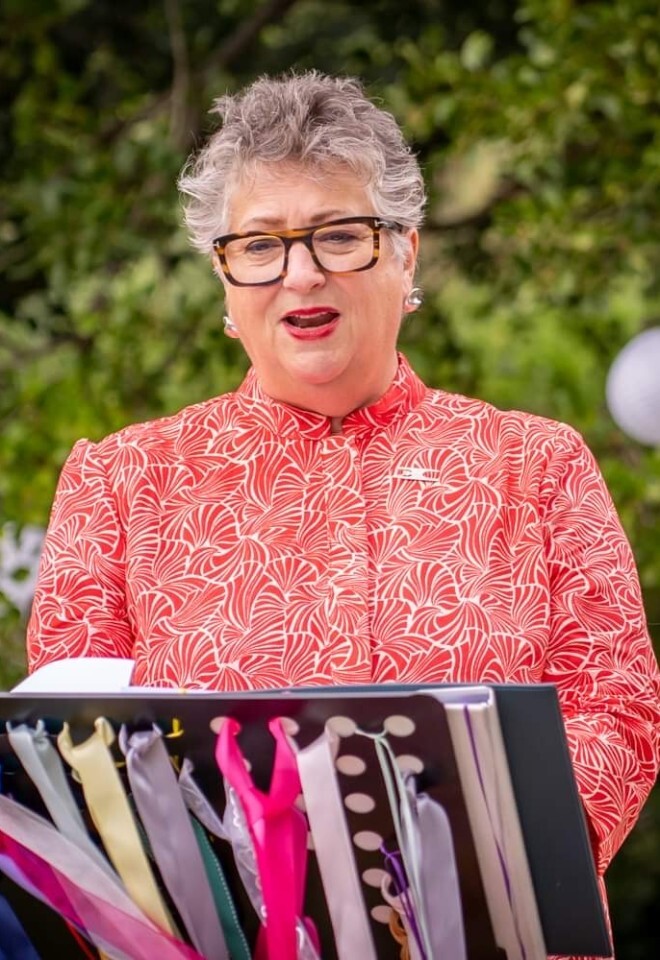 Portrait of celebrant Dawn Rees standing outdoors beside a lectern decorated with colourful handfasting ribbons.