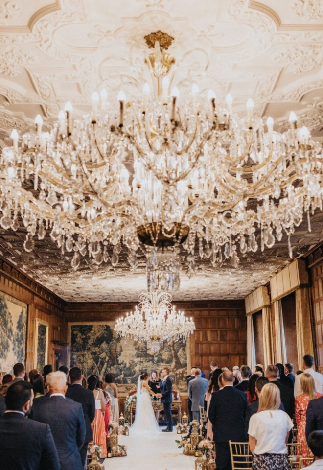 Civil ceremony taking place in the Long Gallery at North Mymms Park, seen from behind the guests under large crystal chandeliers, with ornate plasterwork ceilings and tapestry-covered wooden walls.