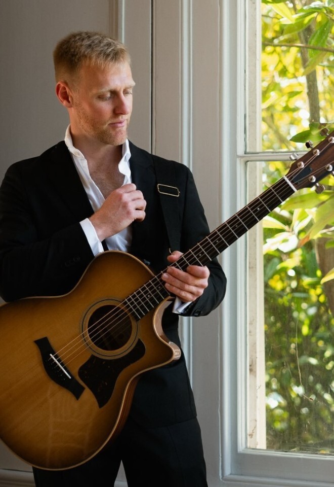 Matt Rayner, an Essex-based wedding singer, stands by a window holding his acoustic guitar, wearing a black suit 