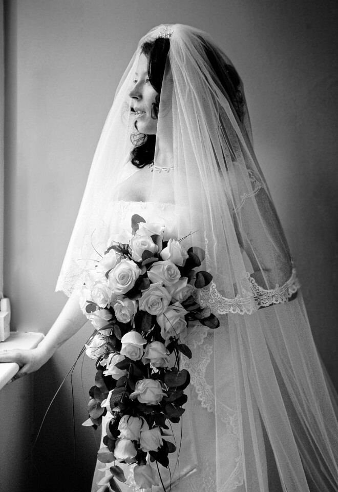 Bride in silhouette standing in a window holding her bouquet, black and white portrait.