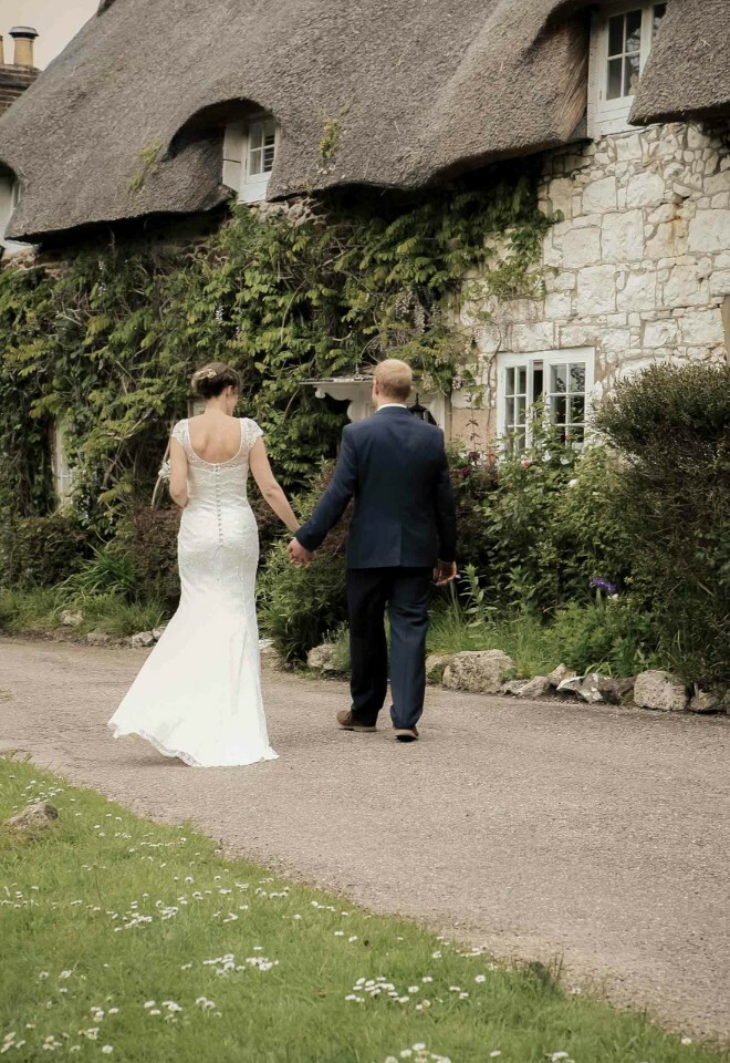 Bride and groom holding hands, walking past a charming thatched cottage.