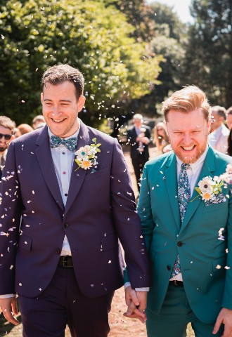 Two grooms smile as their guests thrown confetti over them