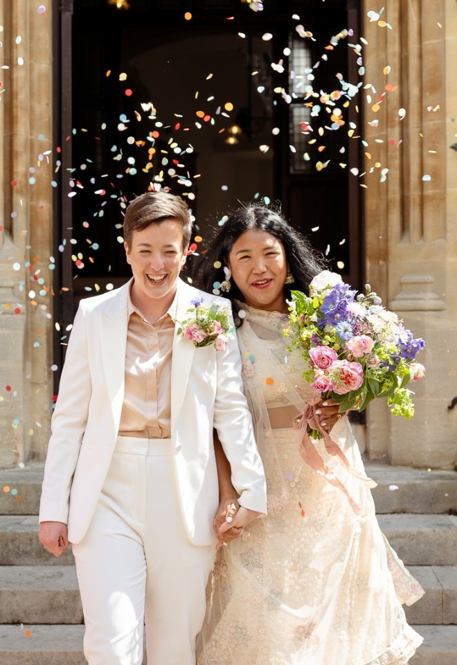 Brides walking under colourful confetti as they exit the registry office, their guests throwing confetti on either side