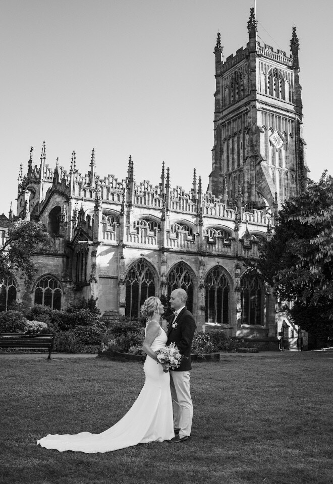 Bride and groom standing together in front of Cirencester Abbey on their wedding day, photographed in black and white.